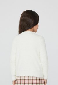 Weißer, flauschiger Pullover mit rundem Halsausschnitt und langen Ärmeln, getragen über einem karierten Rock. Weiche Textur des Kleidungsstücks sichtbar.