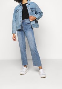 Giacca di denim azzurro chiaro, chiusura con bottoni, due tasche sul petto; abbinata a jeans ampi azzurri chiari con fondo a spacco. Sneakers bianche.