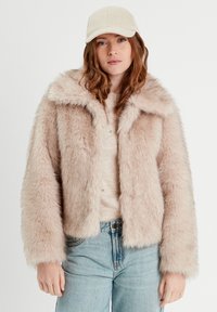 Veste en fausse fourrure rose clair avec un large col et des manches longues. Associée à un pull en tricot beige et à un jean bleu clair.