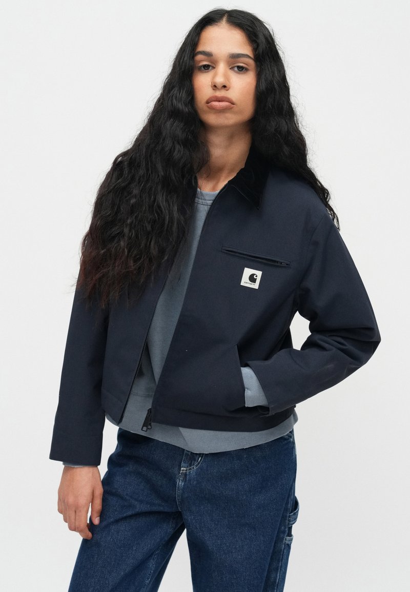 Mørkeblå cropped jakke med lynlås, frontlomme og et logo-mærkat. Har en skjortekrave og struktureret stof.