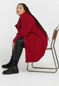 Cappotto in pile rosso con finitura testurizzata, dotato di colletto a punta e dettagli con bottoni. Abbinato a pantaloni in ecopelle neri e stivaletti.