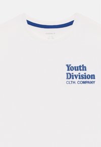 Balts kokvilnas T-kreklis ar apaļu kaklu, kurā kreisajā krūšu daļā ir tumši zils teksts "Youth Division CLTH. COMPANY".