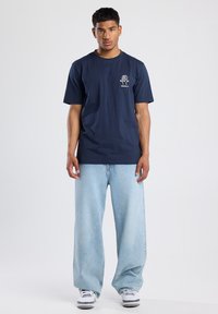 Camiseta de algodón azul marino de manga corta, con un gráfico de un árbol blanco y texto en el lado izquierdo del pecho. Se lleva con unos vaqueros anchos de color azul claro.