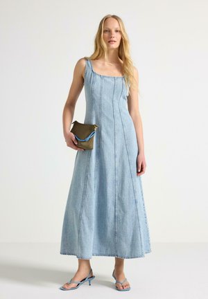 Kvinde iført lyseblå ærmeløs denim maxikjole, der holder en olivengrøn clutch med blå rem, og iført blå stropper på hælene.