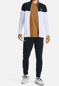 Mans atletiska träningsoverall med en svart och vit zip-jacka och brunt skjorta, ihop med svarta byxor. Materialet är mjukt och lätt.