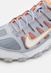 Zapatilla de running Nike en gris y naranja, con malla transpirable, superposiciones sintéticas y suela de goma, mostrada en un primer plano de lado.