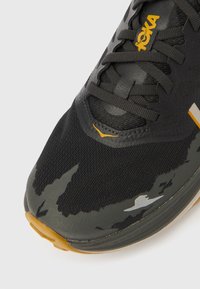 Μαύρο και γκρι αθλητικό παπούτσι με υφασμάτινη επάνω επιφάνεια, που διαθέτει ανακλαστικά στοιχεία και ανθεκτική λαστιχένια σόλα. Κίτρινο λογότυπο στο πλάι.