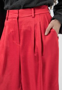 Pantalons plissés rouges avec une texture lisse, taille haute, poches latérales et passants de ceinture, associés à un pull gris foncé.
