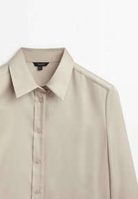 Chemise beige à manches longues avec boutons et col pointu, présentée à plat sur un fond blanc.
