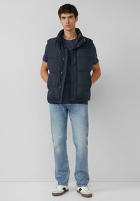Navy blauwe gewatteerde bodywarmer met hoge kraag en drukknoopsluiting, gedragen over een donker T-shirt, gecombineerd met lichtblauwe spijkerbroek en witte sneakers.