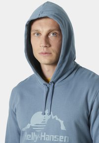 Modrá mikina s kapucí vyrobená z měkké látky, se stylem hor a logem v světle modré barvě. Obsahuje stahovací šňůrky a klokaní kapsu.