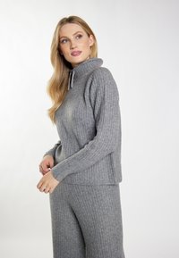 Pulover gri tricotat cu guler înalt și detaliu cu șnur. Are textură canalată și sleeves lungi. Asortat cu pantaloni canalati în aceeași nuanță.