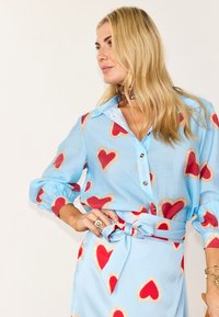 Ξανθιά γυναίκα που φοράει γαλάζιο πουκάμισο με κουμπιά και φούστα με κόκκινα και μπεζ σχέδια καρδιών, ποζάροντας με το χέρι στη μέση μπροστά σε λευκό φόντο.