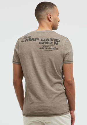 Mann, der mit dem Rücken zur Kamera steht, trägt ein ausgewaschenes braunes kurzärmeliges Hemd mit schwarzem Text am oberen Rücken, auf dem "Camp David Green" und weitere kleinere Schrift zu sehen sind.