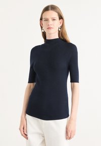 Mørkeblå kortermet strikket topp med høy hals, med ribbestruktur og tettsittende fasong. Merker av sømdetaljer på sidene.