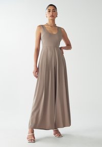 Sansihiheng, jumpsuit taupe dengan leher scoop, desain berlipit, dan kaki lebar. Dipadukan dengan aksesori perak dan sandal tali.