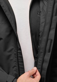 Zwarte jas met een gladde buitenkant en een zachte fleece voering, voorzien van een ritssluiting en klittenbandaccenten. Draagt over een lichtgekleurde shirt.