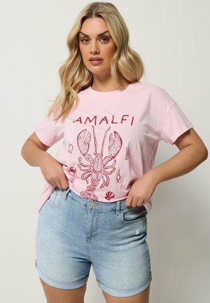 Kvinde med langt blondt hår bærer lyserød t-shirt med rød hummert og teksten "AMALFI", kombineret med lyseblå denimshorts.