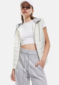 Geheftet Weste in Creme mit grauem Kapuzen, Frontreißverschluss und Seitentaschen, kombiniert mit einem grauen elastischen Taillenbund und einem weißen Cropped-Top.