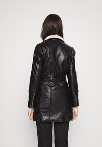 Abrigo de cuero negro con cintura con cinturón, cuello de piel de oveja y cremalleras en las mangas. Textura suave con una silueta ajustada.