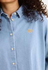 Camicia di jeans azzurro chiaro con chiusura a bottoni, colletto e un piccolo logo a forma di cuore dorato sul petto. Tessuto morbido, vestibilità casual.