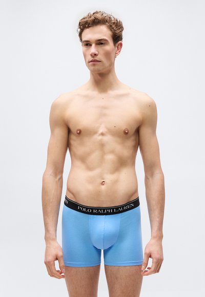 Νεαρός άνδρας με σγουρά μαλλιά, όρθιος χωρίς πουκάμισο, φορώντας ανοιχτό μπλε εσώρουχα boxer Polo Ralph Lauren, σε απλό ανοιχτό φόντο.