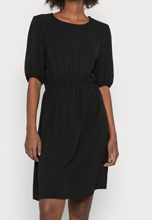 Woman wearing a knee-length black dress with puffed short sleeves and a gathered waist against a plain light background.