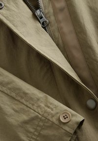 Khakifarbene Jacke mit Reißverschluss, Knopfdetails und einem glatten, strukturierten Stoff. Enthält einen kontrastierenden gewebten Akzent entlang der Naht.