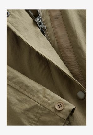 Khaki värviga jakk, mis sisaldab tõmblukku, nööbidetailide ja sileda, tekstuuriga kangaga. Kaasas kontrastne kudumise aktsent õmbluse ääres.