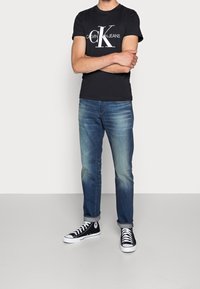 Svart kortärmad T-shirt med vit "Calvin Klein Jeans" logotyp, tillsammans med blå denimjeans och svarta högre sneakers.