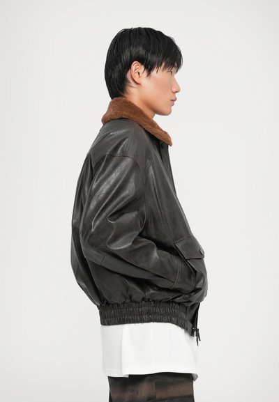 Brown shearling-collared bomber jacket made of dark leather. Features a zip front, elasticized waist, and side pockets. Sleek and oversized fit.