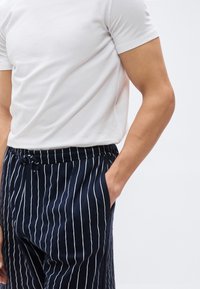 Άνδρας με λευκό κοντομάνικο μπλουζάκι και μπλε σκούρο παντελόνι με ρίγες και κορδόνι στη μέση, το δεξί του χέρι στην τσέπη, με κροπαρισμένη εικόνα από τους ώμους.