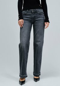 Jeans de mezclilla gris con un ajuste relajado, que cuentan con un diseño clásico de cinco bolsillos, con un sutil desgaste y detalles en el dobladillo deshilachado, combinados con tacones negros.