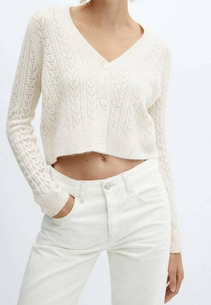 Femme portant un cardigan court en tricot de couleur crème avec des boutons et un jean blanc taille haute, une main dans la poche sur un fond uni.