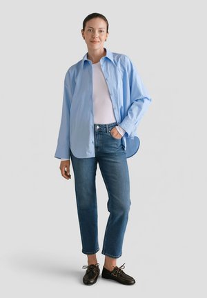 Kobieta stojąca z ręką w kieszeni, ubrana w jasnoniebieską koszulę zapinaną na guziki, białą bluzkę, niebieskie dżinsy i czarne buty na tle białego tła.