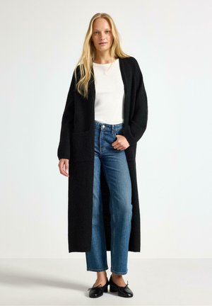 Donna con i capelli biondi lunghi che indossa un lungo cardigan nero, una maglietta bianca, jeans blu e scarpe nere con tacchi bassi, in piedi su sfondo bianco.