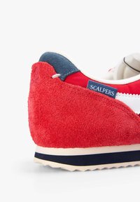 Zapatilla de ante roja con un acento textil azul marino, etiqueta de logo blanca y talón texturizado. Presenta una suela ligera con un patrón en zigzag.