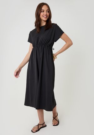 Frau lächelnd, stehend, trägt ein schwarzes Midikleid mit kurzen Ärmeln und einem verstellbaren Taillengürtel, kombiniert mit schwarzen Riemchensandalen.