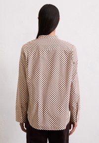Chemise à manches longues en tissu léger, présentant un motif à pois marron sur fond crème, avec un ourlet arrondi et des poignets boutonnés.