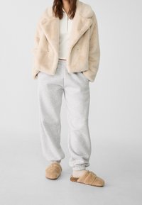 Beiger Kunstpelzmantel, weißes geripptes Top, graue Jogginghose und beigen flauschigen Hausschuhe. Weiche Texturen und entspannte Passform im gesamten Outfit.