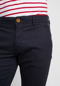 Granatowe bawełniane spodnie mają zapięcie na guzik z jasnobrązowym akcentem, szczegółowe szwy i slim fit, stanowią część codziennego stroju.