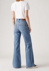 Helle blaue Schlagjeans mit hoher Taille, fünf Taschen und einem braunen Lederpatch auf der Rückseite, kombiniert mit einem weißen T-Shirt und schwarzen Sneakers.