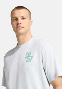 Harmaa puuvillainen T-paita, jossa vihreä graafinen kuvio ja numerot "1957". Lyhyet hihat ja klassinen pyöreä pääntie.