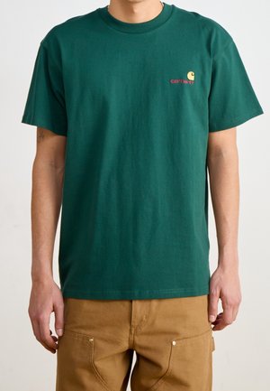 Uomo che indossa una t-shirt Carhartt a maniche corte verde scuro e pantaloni marrone chiaro, in piedi con le braccia rilassate lungo i fianchi.