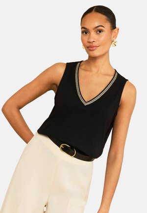 Žena nosící černý top bez rukávů s výstřihem do V a zlatým lemováním, krémové vysoké kalhoty, černý pásek se zlatou přezkou a zlaté náušnice.