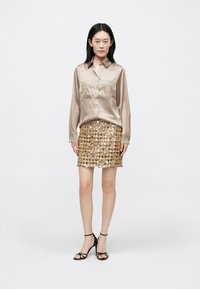 Chemise beige satinée avec fermeture à boutons, poches poitrine, associée à une mini-jupe en sequins dorés et des sandales noires à brides.