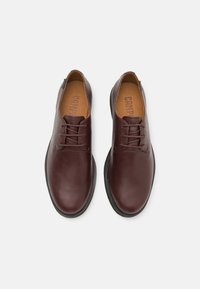 Scarpe in pelle marrone con texture liscia, punta tonda e sistema di allacciatura. Presentano una suola in gomma nera e una soletta color tan con branding.