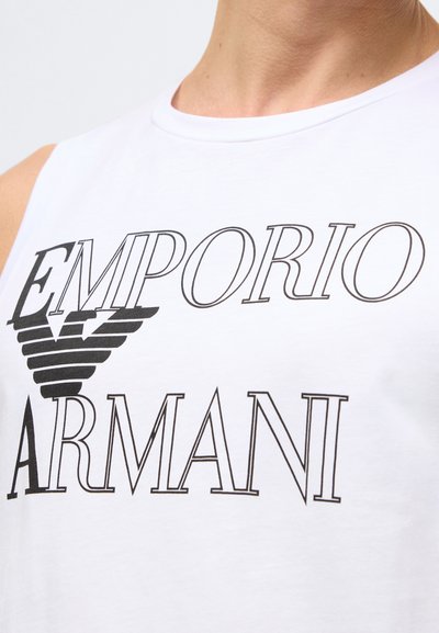 Biały bawełniany top na ramiączkach z nadrukiem "EMPORIO ARMANI" w czerni oraz stylizowanym logo orła na środku, minimalistyczny design.