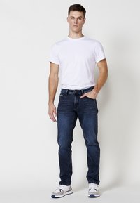 Jeanșii bărbați din denim, de culoare albastru închis, cu croială slim, design cu cinci buzunare și detaliu brodat pe buzunarul din spate, asortați cu un tricou alb simplu.