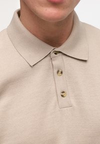 Beige polo skjorte med ribbet tekstur, foldet krave og tre gyldne knapper foran, som viser en uformel design.